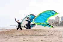 Cheerful multiethnic female kiters with power kites jumping above sandy ocean coast under white sky in city — Stock Photo
