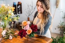 Florista femenina joven concentrada con el pelo ondulado largo en ropa casual y delantal que arregla el ramo elegante de rosas y flores surtidos en la tienda - foto de stock