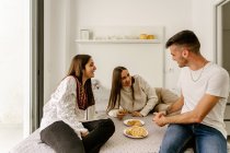 Cheerful young ethnic male and female friends in casual clothes relaxing on bed and eating yummy pastries during weekend at home — Foto stock