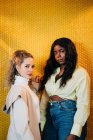 Selbstbewusste multiethnische Frauen in trendiger Kleidung stehen zusammen auf gelbem Hintergrund und blicken in die Kamera — Stockfoto