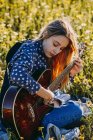 Giovane donna hipster riflessiva seduta su un prato in campagna a suonare la chitarra durante la luce del sole estiva — Foto stock
