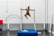 Full body muscular strong sportsman in shorts performing exercise on aerial silks in modern light fitness center — Stock Photo
