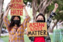 Mujeres étnicas con máscaras y con Stop Asian Hate y Asian Lives Matter afiches protestando contra el racismo en la calle de la ciudad y mirando a la cámara - foto de stock