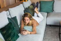Mulher alegre deitada no sofá e desfrutando de música em fones de ouvido enquanto olha para a tela do smartphone — Fotografia de Stock