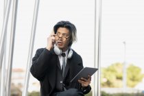 Молодой предприниматель-этнический мужчина в современных наушниках и очках с планшетом, смотрящим в город — стоковое фото