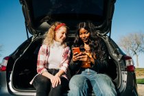 Baixo ângulo de amigos femininos multirraciais alegres sentados no porta-malas do carro e navegando nas mídias sociais no telefone celular juntos — Fotografia de Stock