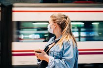 Junge Frau in Freizeitkleidung und Schutzmaske steht auf Bahnhof und schaut weg, während sie auf Zug wartet — Stockfoto