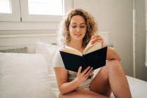 Mujer joven positiva con pelo rubio rizado en bragas y anteojos sonriendo mientras está sentada en una cama acogedora y leyendo un libro interesante - foto de stock