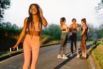 Cheerful African American female athlete listening to music in headphones while standing in park at sunset on background of company of blurred sportswomen — Stock Photo