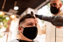 From below stylist in textile mask with hair dryer against man in cape in armchair in barbershop — Stock Photo