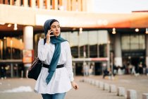 Этническая женщина в хиджабе, стоящая рядом с железнодорожным вокзалом и разговаривающая по мобильному телефону, отворачиваясь — стоковое фото