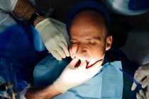 Crop anonymous orthodontist in latex gloves with coworker checking up teeth of mature man looking forward in hospital — Stock Photo