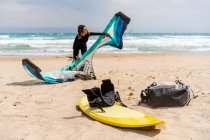 Female kiter in wetsuit setting up inflatable kite on sandy ocean coast with backpack and harness on kiteboard — Stock Photo