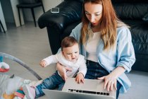 Junge Mutter in lässiger Kleidung umarmt niedliches Baby und blättert im Netbook, während sie zusammen auf dem Boden sitzt — Stockfoto
