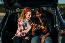 Щасливі багаторасові друзі, що сидять у багажнику автомобіля та переглядають соціальні мережі на мобільному телефоні разом — стокове фото
