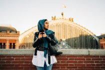 Muslim female in traditional headscarf standing on city street and browsing phone while looking away — Stock Photo