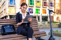 Content young ethnic female entrepreneur sitting on bench while browsing on tablet near electric scooter and city buildings — Stock Photo