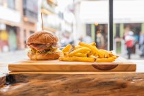 Вкусный свежий гамбургер и хрустящая картошка фри подаются на деревянной доске в современном ресторане быстрого питания — стоковое фото