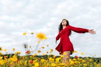 From below side view of trendy female in red sundress and with flower crown standing with eyes closed on blossoming field with yellow and red flowers with outstretched arms on warm summer day — Stock Photo