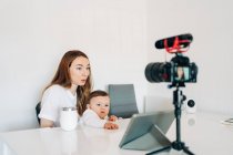 Joven madre con un lindo bebé en vueltas hablando y grabando video en la cámara para blog personal mientras está sentado en el escritorio en casa - foto de stock