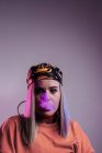 Cool female in street style outfit smoking e cigarette and exhaling smoke through nose on purple background in studio with pink neon illumination — Stock Photo