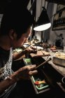 Vue latérale d'un artisan ethnique regardant de près une pièce métallique et frappant un timbre métallique avec un petit marteau — Photo de stock