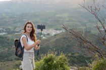 Side view of traveling female with backpack standing on hill and taking self shot on smartphone on background of mountain range in summer — Stock Photo