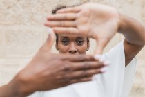 Fröhliche junge Afroamerikanerin in weißer Bluse, die auf Kameraschildern mit Händen zeigt und in die Kamera blickt, während sie vor einer unebenen Wand steht — Stockfoto