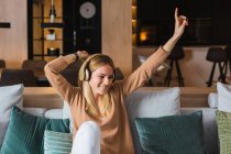 Optimistische Frau sitzt auf Sofa und hört Musik über Kopfhörer, während sie Lieder mit erhobenen Händen genießt — Stockfoto