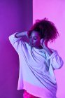 Giovane donna afroamericana in abiti bianchi che indossa cuffie sul collo mentre in piedi con gli occhi chiusi in studio buio in luci al neon — Foto stock