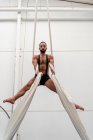 From below full body muscular strong sportsman in shorts performing exercise on aerial silks in modern light fitness center — Stock Photo