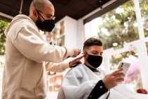 Von unten Stylist mit Trimmer schneiden Haare des Mannes in Stoff Gesichtsmaske mit Handy im Friseurladen — Stockfoto