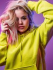 Donna determinata in felpa gialla con cappuccio che tocca i capelli biondi in piedi su sfondo viola in studio con luci al neon blu e rosa — Foto stock