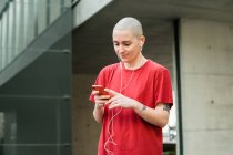 Joven mujer homosexual feliz en camiseta y auriculares con teléfono celular mirando a la pantalla mientras escucha música - foto de stock