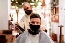 Male hairdresser in cloth face mask with brush removing hair from cape of client in barbershop — Foto stock