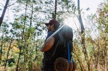 Dal basso barbuto zaino in spalla maschile in berretto passeggiando tra alberi e piante nei boschi nella giornata di sole — Foto stock