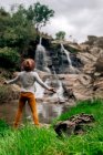 Indietro vista anonimo zaino in spalla maschile in cappello godendo vista di cascata in streaming da roccia grezza nella natura verdeggiante — Foto stock