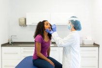 Especialista médica femenina en uniforme protector, guantes de látex y mascarilla para la cara haciendo una prueba de hisopo bucal con brote de algodón en una paciente afroamericana en una clínica durante el brote de coronavirus - foto de stock