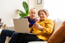 Cheerful mature couple making payment with plastic card during online shopping via laptop at home — Stock Photo