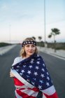 Encantada americana fêmea de pé envolto com bandeira nacional dos EUA na estrada ao pôr do sol e olhando para longe — Fotografia de Stock