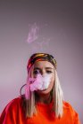 Прохолодна жінка в наряді вуличного стилю курить електронну сигарету і видихає дим через ніс на фіолетовому фоні в студії з рожевим неоновим освітленням — стокове фото