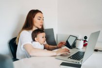 Focused young mother working on laptop holding curious baby watching funny video on tablet while sitting together at desk in light room — Stock Photo