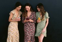 Zufriedene junge beste Freundinnen in trendiger Kleidung mit Mobiltelefonen, die auf dem städtischen Gehweg gegen die Wand stehen — Stockfoto