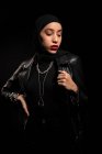 Atractiva joven mujer islámica vestida con traje negro con chaqueta de cuero y hijab mirando suavemente hacia abajo en el estudio negro - foto de stock
