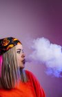 Seitenansicht einer coolen Frau im Streetstyle-Outfit, die E-Zigarette raucht und Rauch durch die Nase auf lila Hintergrund im Studio mit rosa Neonbeleuchtung ausatmet — Stockfoto