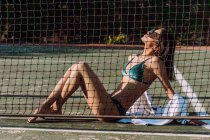 Вид збоку молодої ніжної жінки-туристки в купальниках і сонцезахисних окулярах, що сидять з схрещеними ногами за огорожею сітки під час відпустки — стокове фото