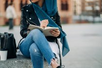 Cultiver femme musulmane dans le hijab écrit dans un journal tandis que assis sur la rue de la ville et regardant loin — Photo de stock
