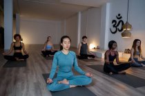 Mulher asiática com grupo de diversas pessoas sentadas em Lotus posar com os olhos fechados e mediar enquanto praticam ioga juntos durante a aula em estúdio — Fotografia de Stock