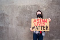 Mujer étnica en máscara y con cartel de cartón con inscripción Asian Lives Matter protestando en la calle de la ciudad y mirando a la cámara - foto de stock