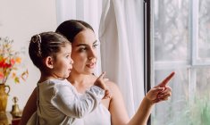 Side view of ethnic mother with cute little daughter looking out of window while standing in room at home — Stock Photo